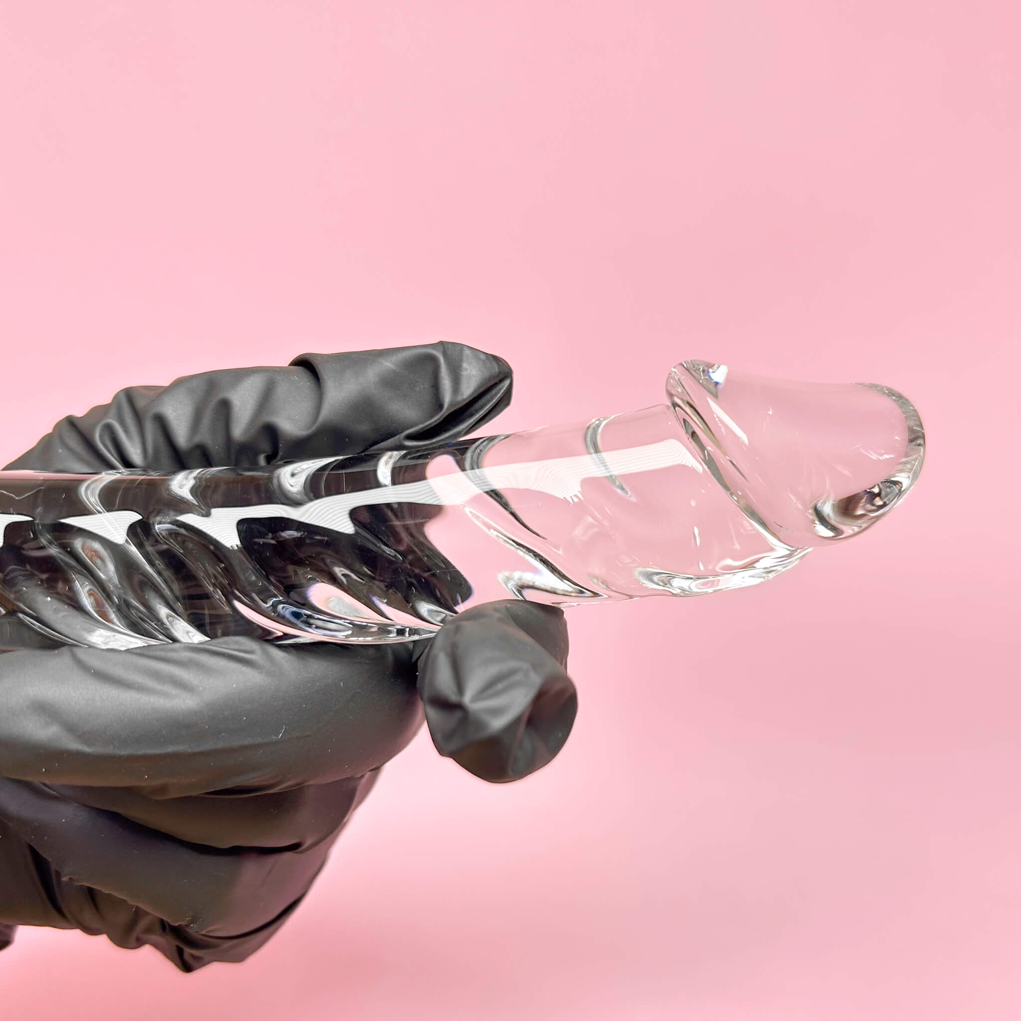 Clear glass dildo held by a gloved hand against a pink background