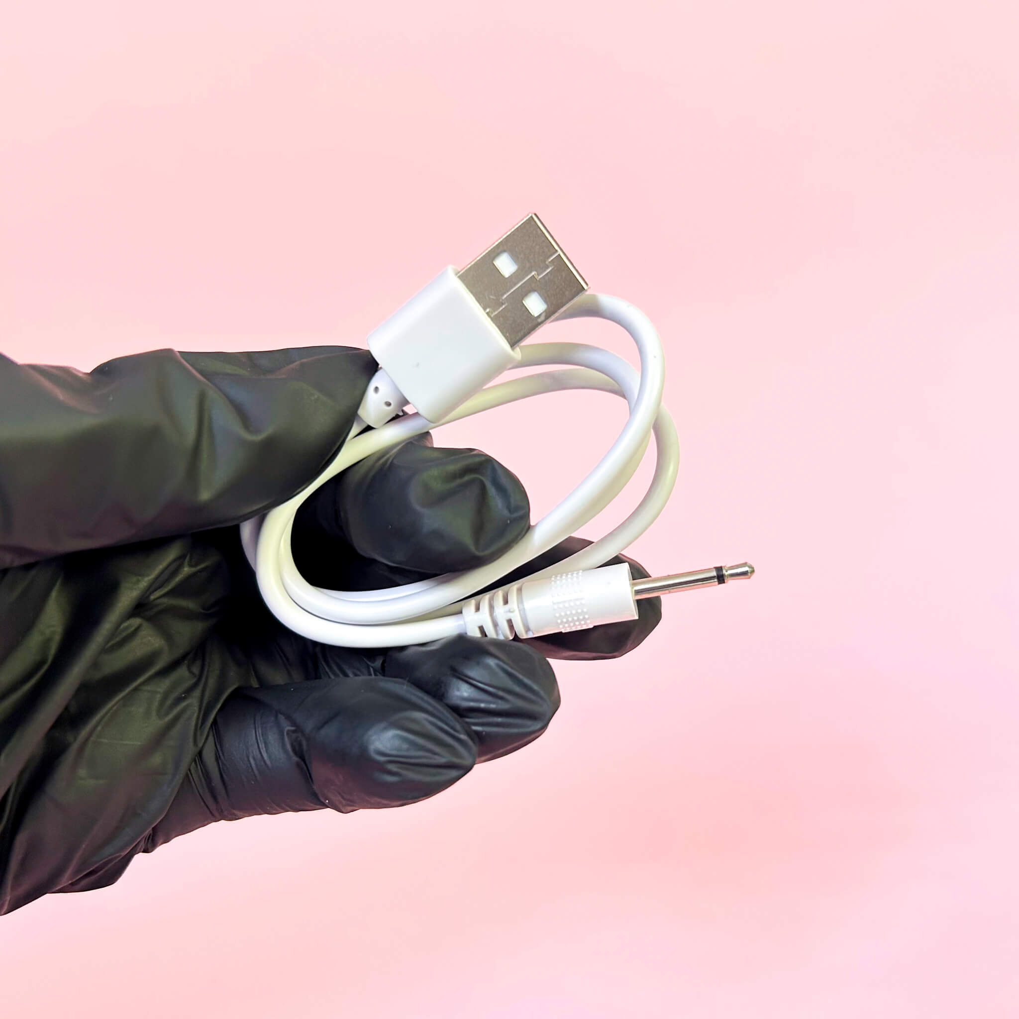 Hand wearing a black glove holding a white USB cable with a 3.5mm plug against a pink gradient background