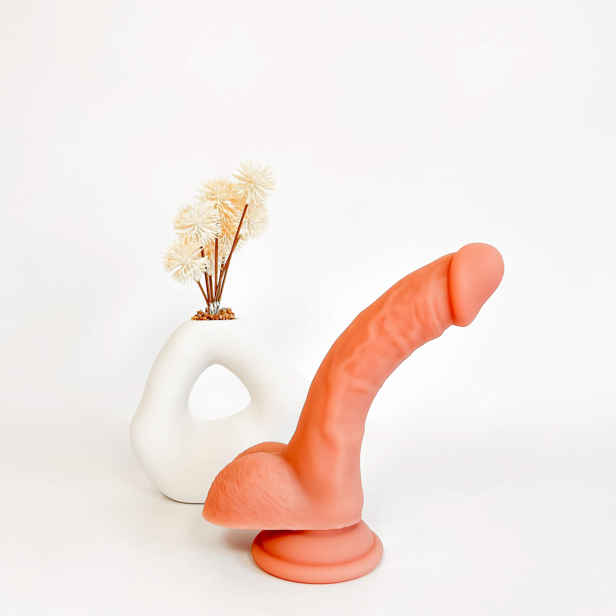 A realistic peach-colored dildo with a textured shaft and flared base, displayed in front of a white sculptural vase holding dried cream flowers on a clean white background.