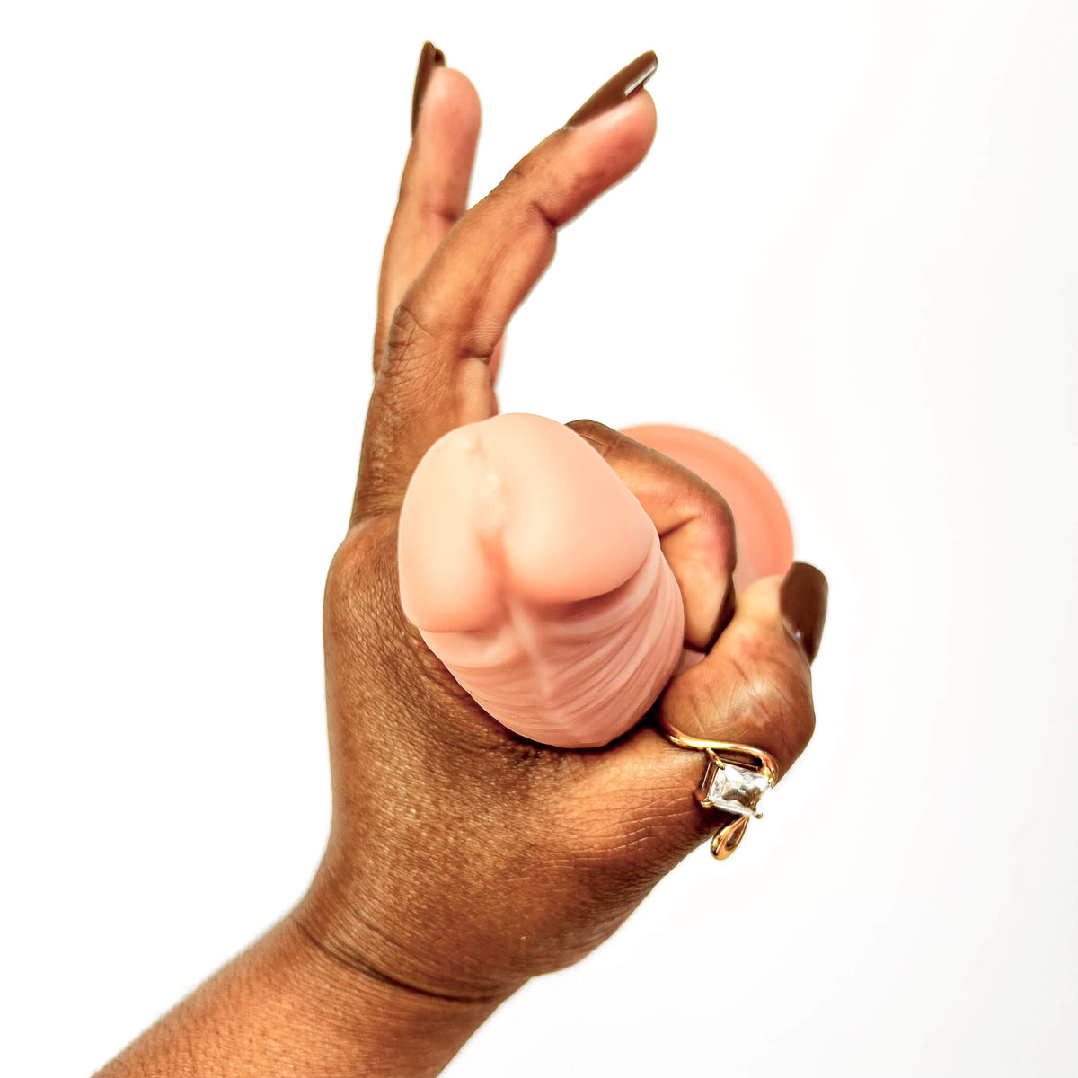 A close-up of a hand holding a realistic silicone dildo against a plain white background. The hand has long brown manicured nails and a gold ring on the ring finger.