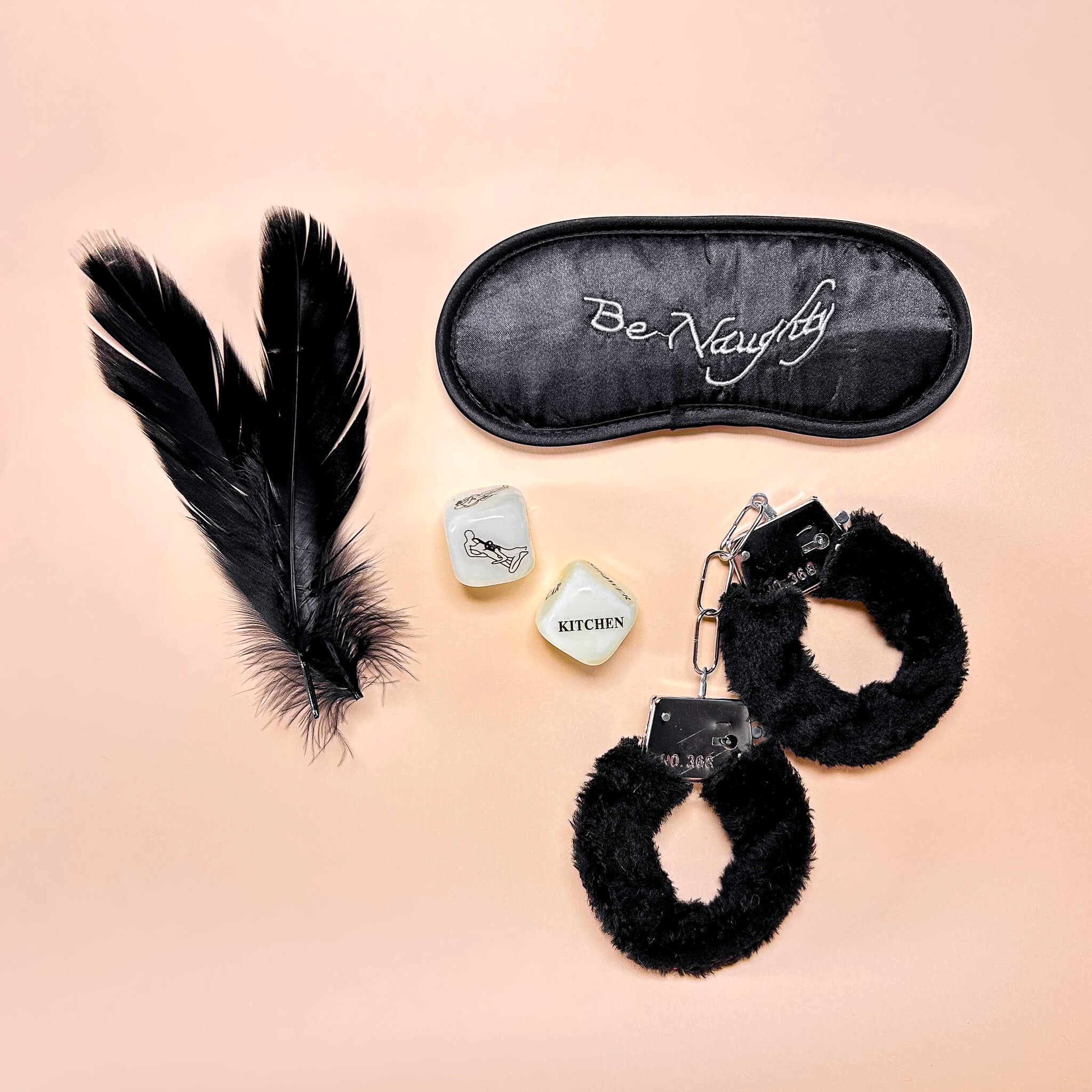 Black feather, sleep mask with 'Be Naughty' text, Dice, and handcuffs on a pink background