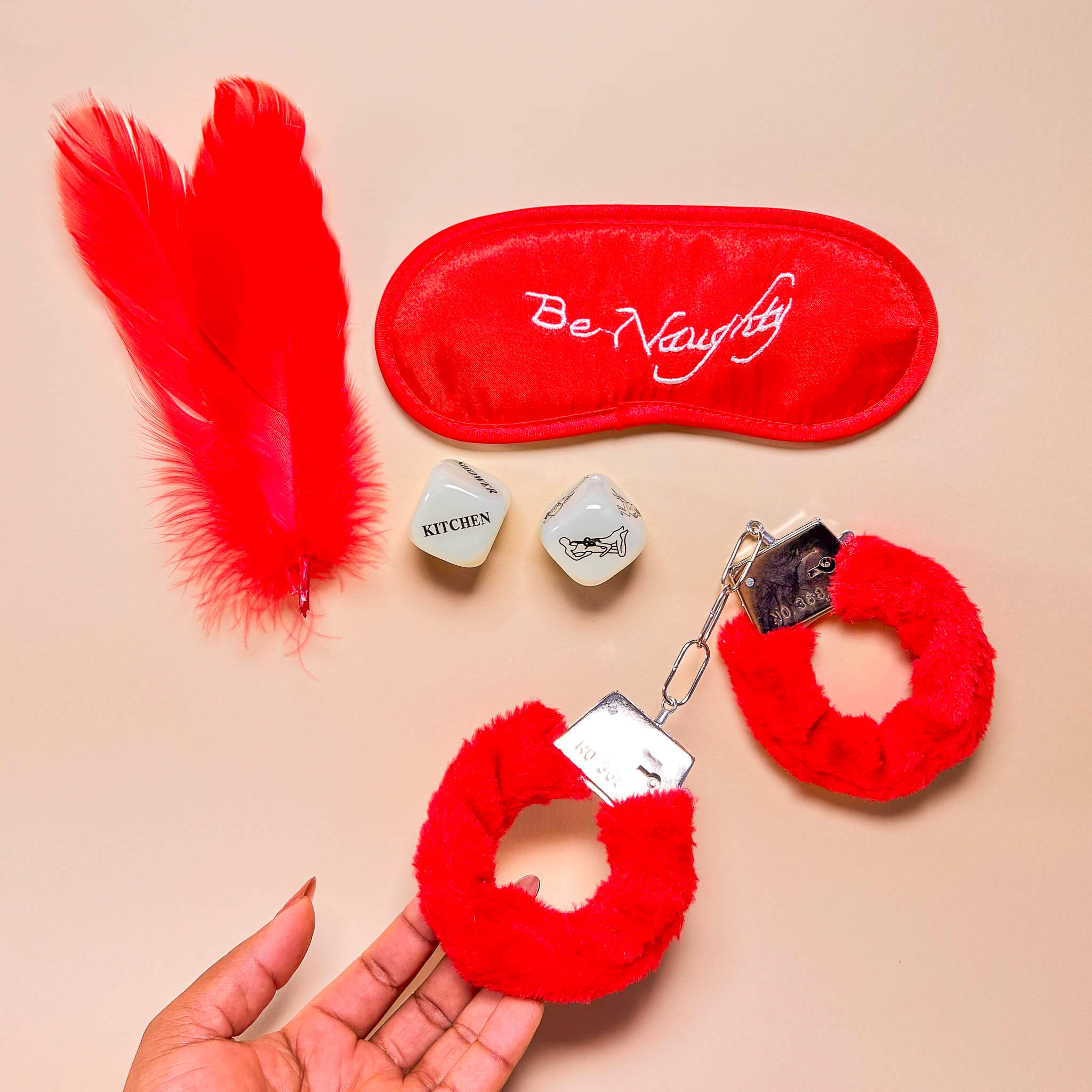 Red feather, 'Be Naughty' eye mask, red fluffy handcuffs, and small adult dice on a beige background.