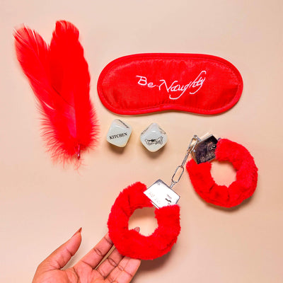 Red feather, 'Be Naughty' eye mask, red fluffy handcuffs, and small adult dice on a beige background.