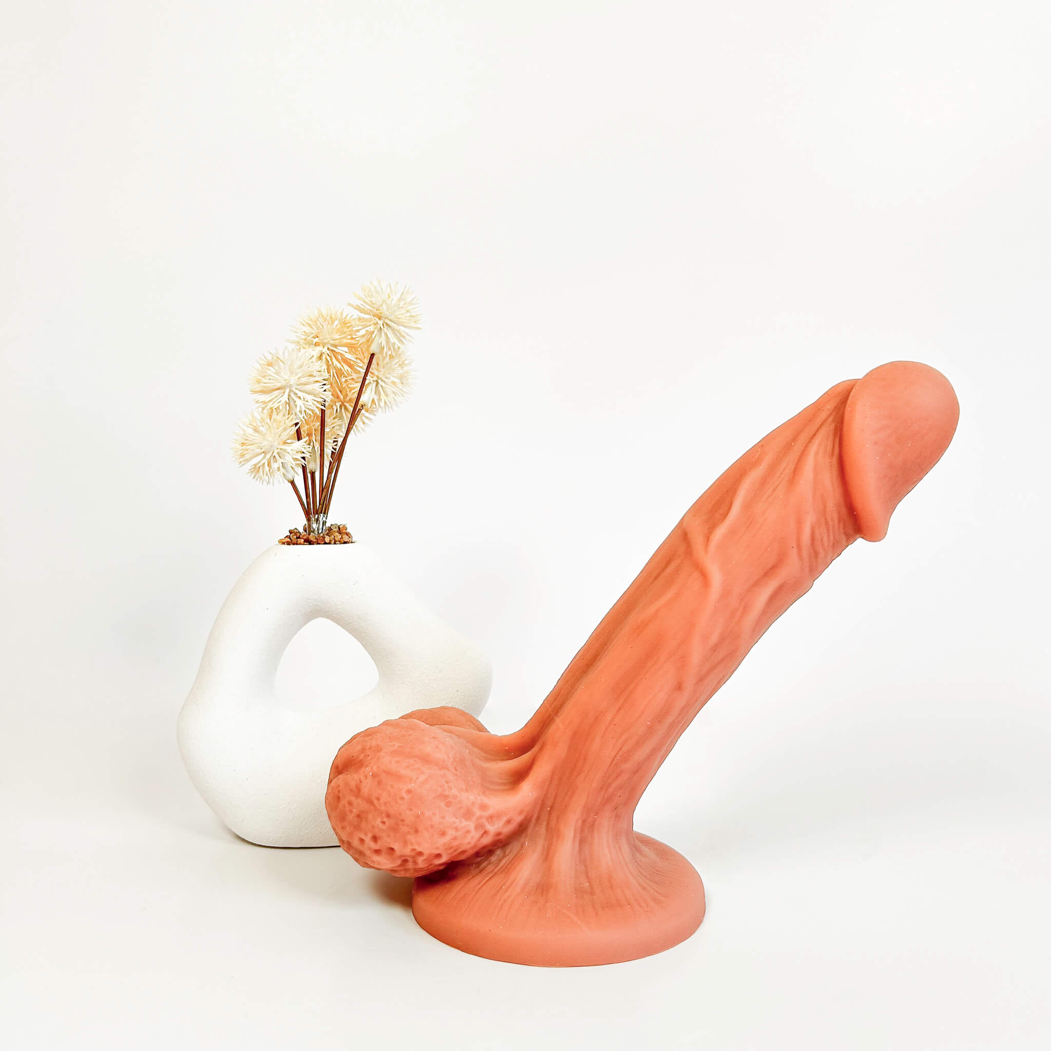 BendBoss Realistic Suction Cup Dildo in a flesh color, styled in front of a white sculptural vase with dried flowers on a seamless white background.
