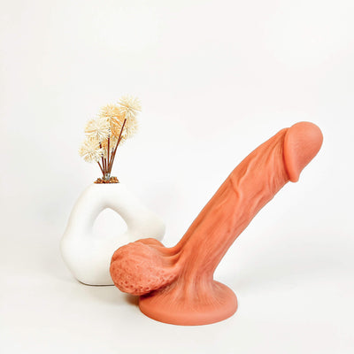 BendBoss Realistic Suction Cup Dildo in a flesh color, styled in front of a white sculptural vase with dried flowers on a seamless white background.