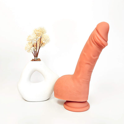 An adult novelty product shaped like a realistic peach-colored dildo standing upright on a round base, photographed on a clean white studio background next to a white sculptural vase with dried stems.