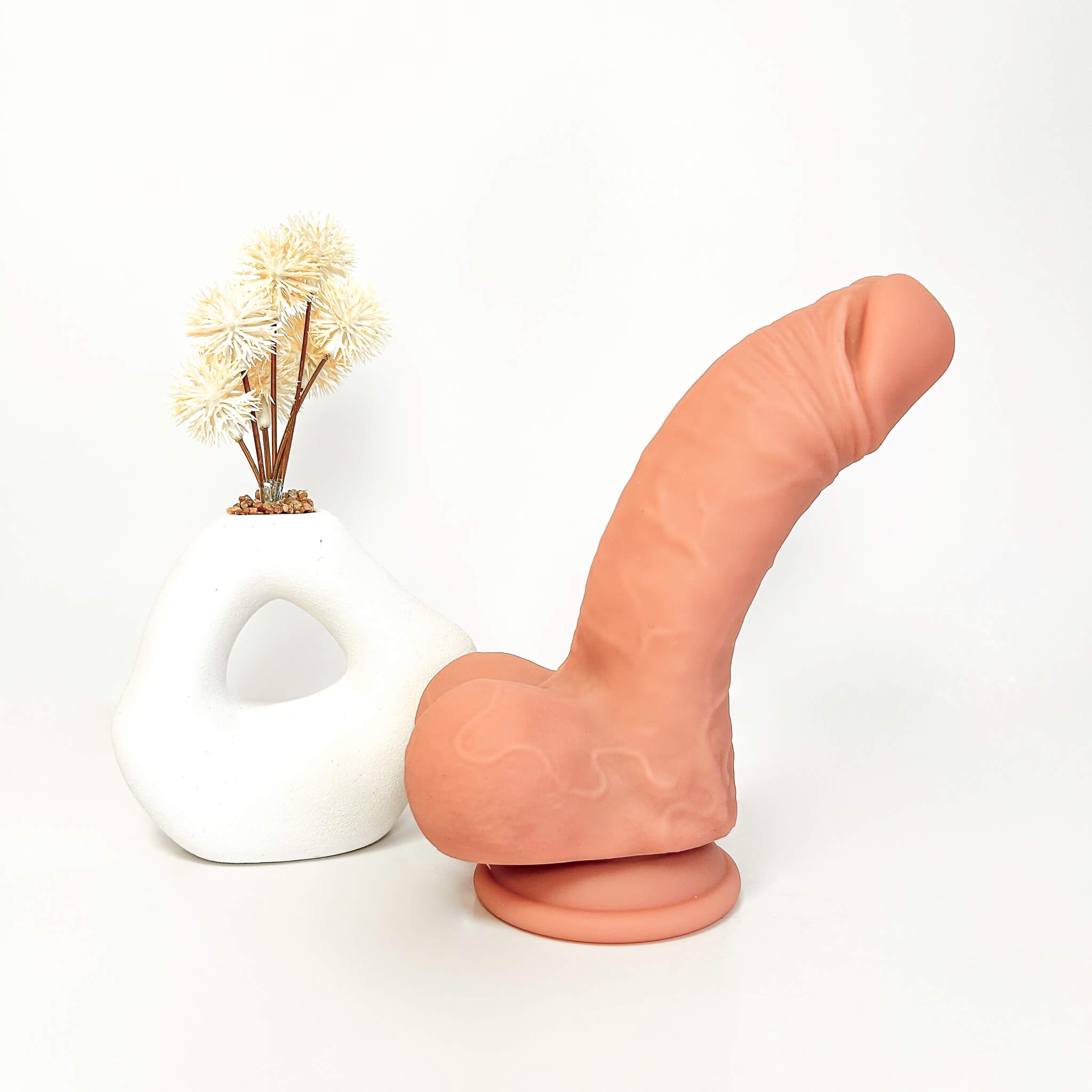 A realistic peach-colored dildo standing upright on a round base next to a white ceramic vase with dried flowers, set against a plain white background.