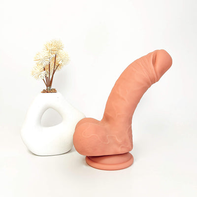 A realistic peach-colored dildo standing upright on a round base next to a white ceramic vase with dried flowers, set against a plain white background.