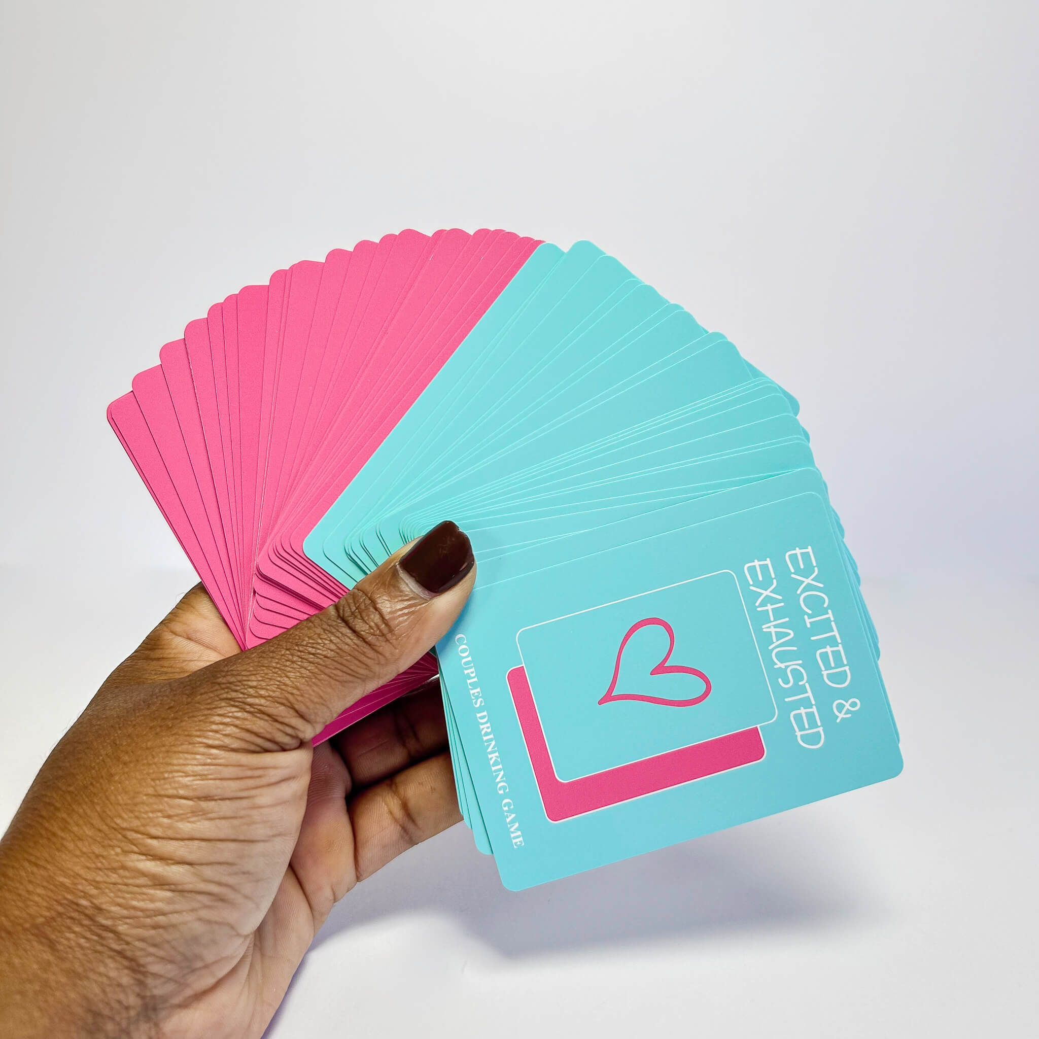 Hand holding pink and blue Excited & Exhausted Couples Game cards with a heart design on a white background