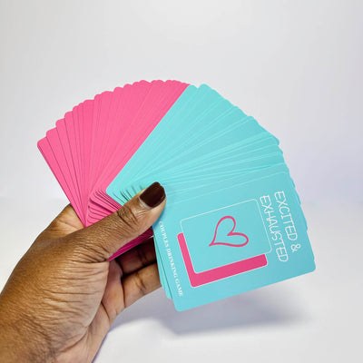 Hand holding pink and blue Excited & Exhausted Couples Game cards with a heart design on a white background