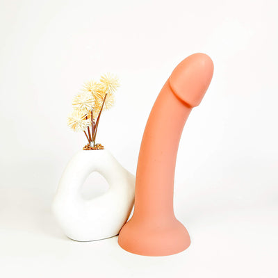 A peach-colored curved silicone dildo standing upright next to a white sculptural vase with dried yellow flowers, set against a plain white background.