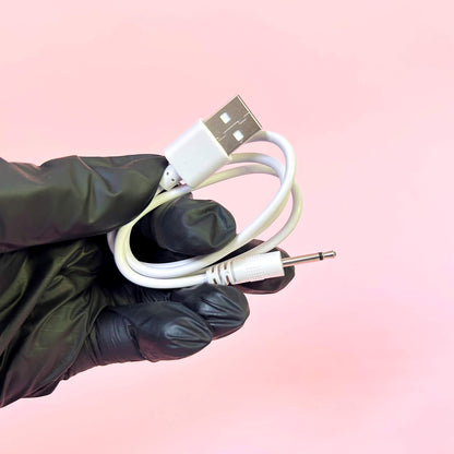 Hand wearing a black glove holding a white USB cable with a 3.5mm plug against a pink gradient background