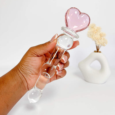 Hand holding a clear glass dildo with a pink heart-shaped stopper on a white background