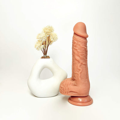 A realistic flesh-toned dildo standing upright next to a small white sculptural vase with dried beige flowers, photographed on a clean white studio background.