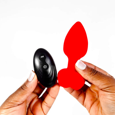 Black remote control and red red remote butt plug held by hands against a white background