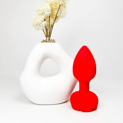 Red silicone butt plug next to a white vase with dried flowers on a white background