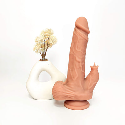 A novelty silicone dildo in a matte peach-pink color stands next to a white sculptural vase with dried cream flowers on a seamless white background.