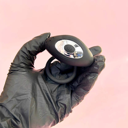 Hand wearing a black glove holding a small vibrating cock ring device against a pink background