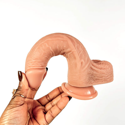 A flesh-toned realistic dildo with a suction-cup base, held in a person's hand against a plain white background.