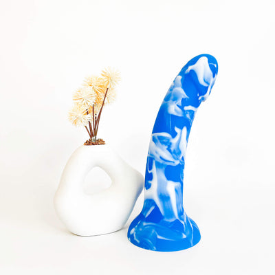 A novelty silicone dildo with a blue and white marbled pattern, displayed next to a white ceramic vase holding dried yellow flowers on a clean white background.