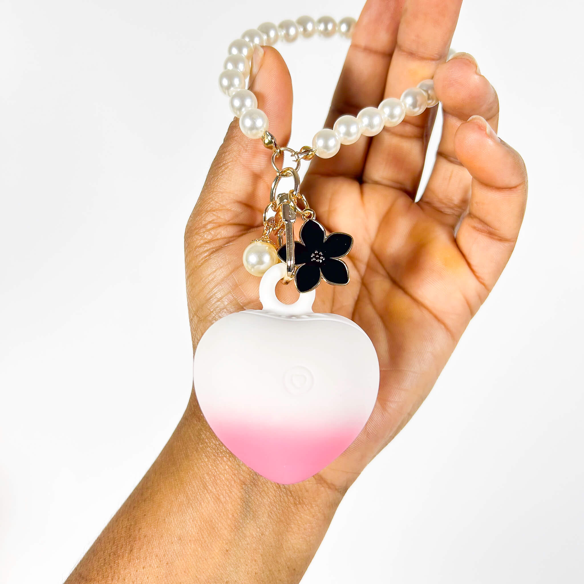 Hand holding a white and pink heart-shaped discreet vibrator for women with a pearl necklace and black flower charm.