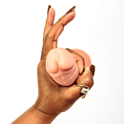 A close-up of a hand holding a realistic silicone dildo against a plain white background. The hand has long brown manicured nails and a gold ring on the ring finger.