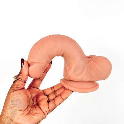 A flesh-toned curved silicone adult novelty item being held in a person's hand against a plain white background.