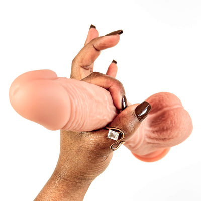 A close-up of a hand with dark brown manicured nails holding two realistic flesh-toned dildo props against a plain white background.