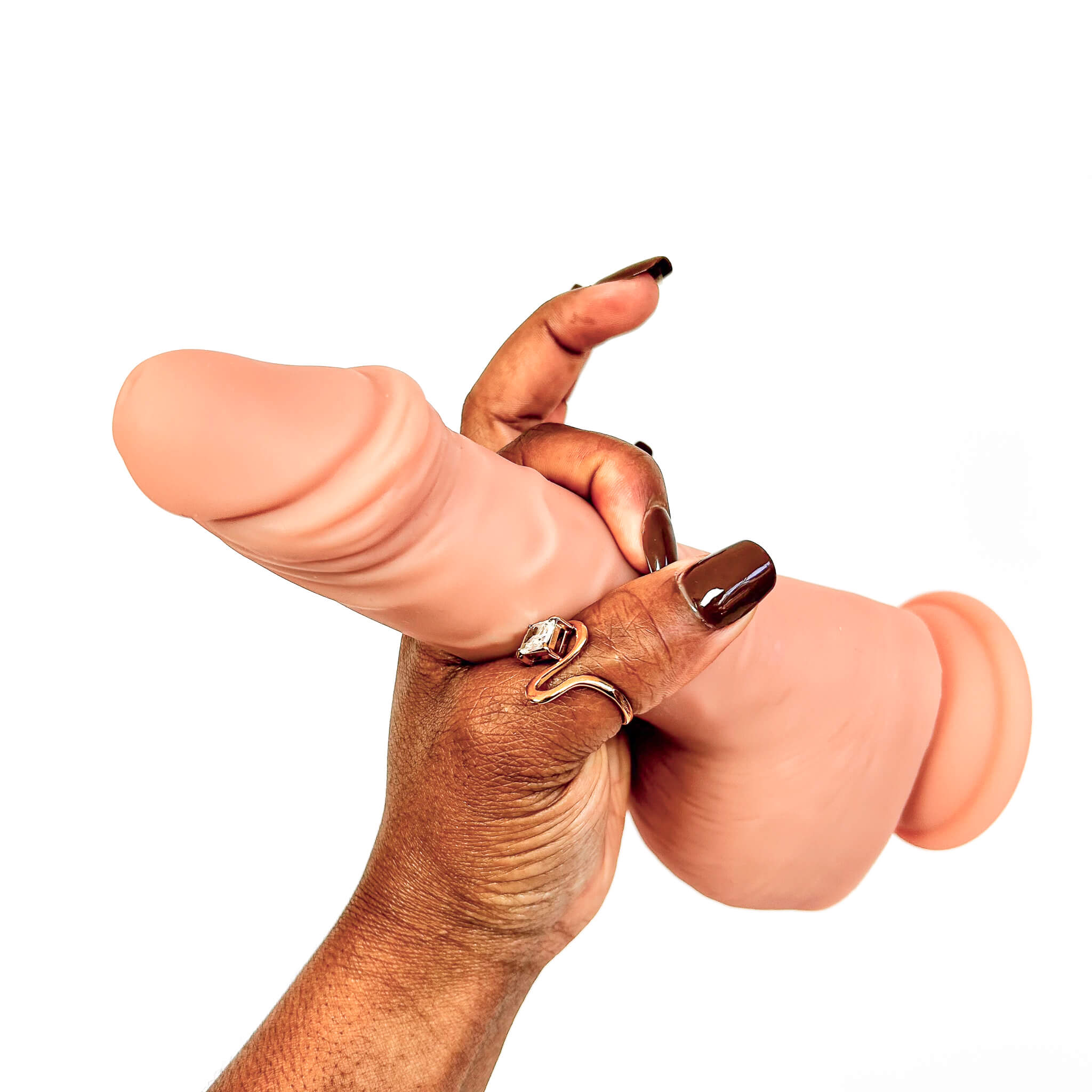 A close-up of a person's hand with dark brown manicured nails and silver rings, holding a light peach-colored silicone adult novelty toy against a plain white background.