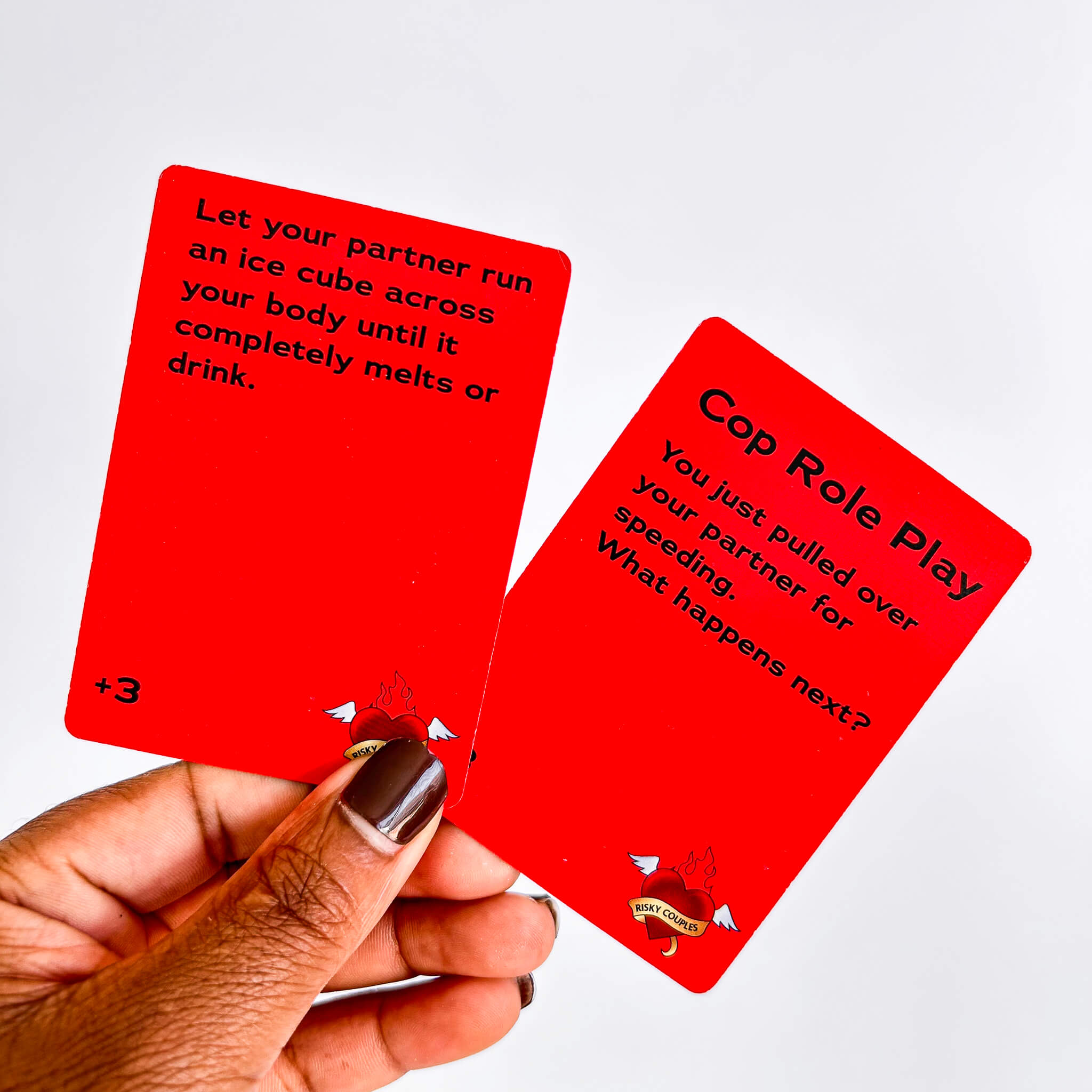 Two red risky couples game cards with text held by a hand against a white background
