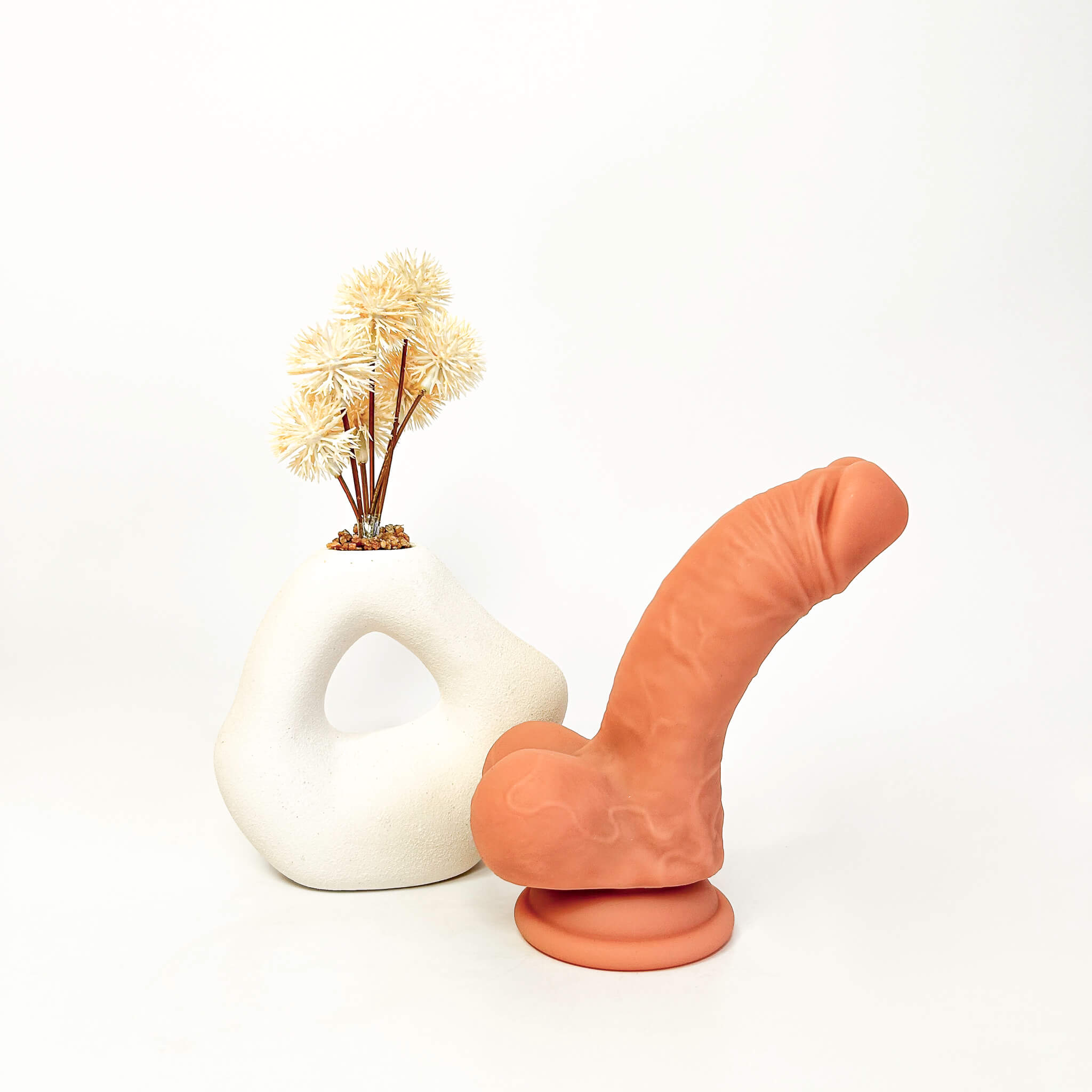 An adult novelty product shaped like a stylized penis in a matte terracotta orange color, displayed next to a white sculptural ceramic vase holding dried grass stems on a seamless white background.