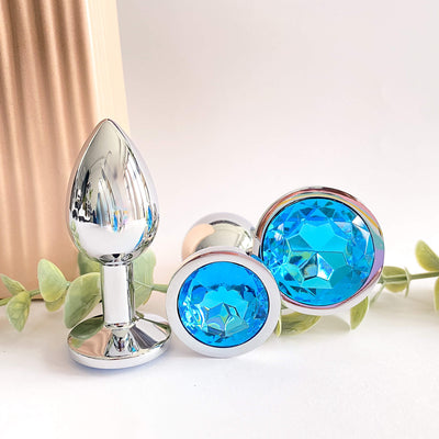 The image displays a set of three light blue metal butt plugs, each featuring a radiant acrylic gem base. The plugs come in different sizes, showcasing a shiny, smooth metal surface that highlights their elegance and sophistication.
